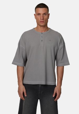 Uomo che indossa una maglia henley a maniche corte grigio chiaro in tessuto a cialda con tre bottoni e pantaloni in denim grigio scuro, in piedi davanti a uno sfondo neutro.