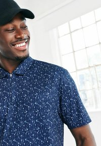 Lyhythihainen tummansininen polo-paita, jossa on valkoinen kuvio erilaisista pienistä esineistä, ja vetoketjukaulus. Materiaali näyttää sileältä ja kevyeltä.