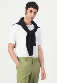 Camiseta blanca con mangas enrolladas, combinada con pantalones en un tono verde claro. Un suéter azul marino atado alrededor de los hombros añade contraste y textura.