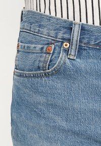 Džíny z denimové látky v světle modré barvě s vybledlým povrchem. Mezi hlavní rysy patří měděné knoflíky a švy, design s pěti kapsami a pásek.