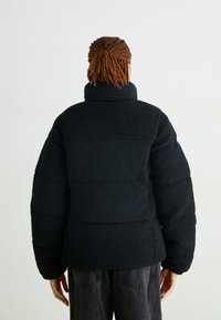 Person med flätat hår iklädd en svart texturerad dunjacka, som står vänd bort mot en enfärgad vit bakgrund.