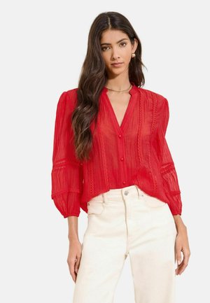 Woman wearing a sheer red long-sleeve blouse with lace details and cream high-waisted pants, standing against a plain background.