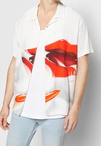 Camisa de manga corta y cuello en tejido blanco con un gran gráfico de labios rojos y lápiz labial, con botones transparentes y un corte relajado.