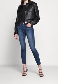 Persona con chaqueta de cuero corta negra, top negro, jeans skinny azules y sandalias negras de tacón alto de tiras, de pie contra un fondo blanco.