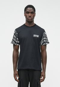 Muž, ktorý má na sebe čierne tričko Versace Jeans Couture s bielym logom a opakovaným textom na rukávoch.