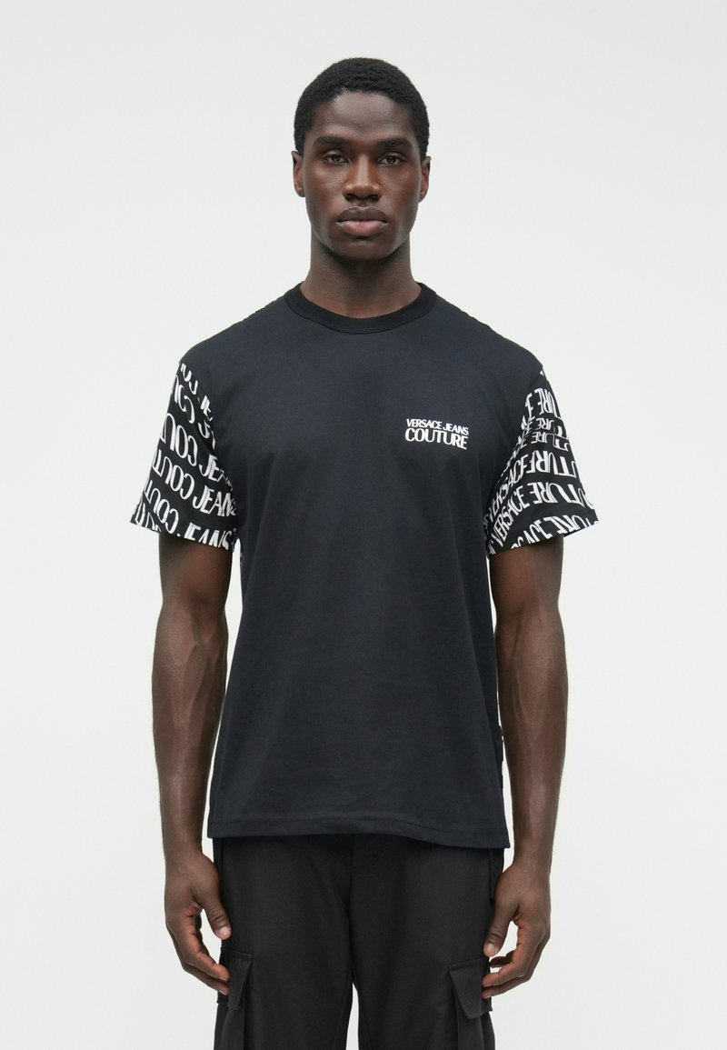 Muž, ktorý má na sebe čierne tričko Versace Jeans Couture s bielym logom a opakovaným textom na rukávoch.