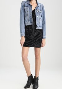 Denim jacket in light blue, cropped design with button closure and two chest pockets. Worn over a black velvet dress, knee-length. Black ankle boots.