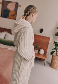 Peignoir en peluche blanche texturée avec des rayures verticales, capuche attachée et poches avant. Tissu doux, porté sur une housse de couette rose pâle.