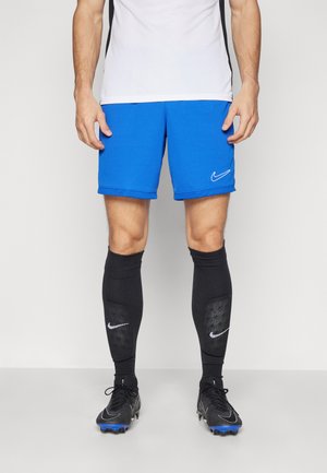 Mėlynos sportinės šortai su baltu Nike logotipu, derinami su juodais iki kelių siekiančiais kojinėmis, turinčiomis tekstūruotus akcentus, ir juodais futbolo bateliais.