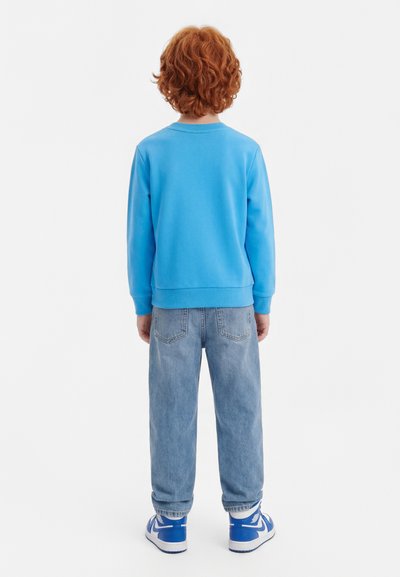 Παιδί με σγουρά κόκκινα μαλλιά στέκεται με την πλάτη γυρισμένη φορώντας μπλε φούτερ, ανοιχτόχρωμα τζιν και μπλε-άσπρα αθλητικά παπούτσια.