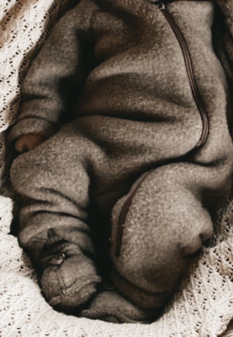 Bébé endormi portant une grenouillère en polaire grise confortable avec pieds, allongé sur une couverture blanche tricotée.