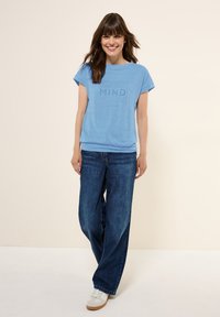 T-shirt à manches courtes bleu clair arborant le texte "Esprit Reconnaissant" dans une teinte légèrement plus foncée. Associé à un jean bleu foncé ample.