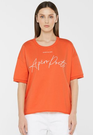 Young woman with straight brown hair wears an orange oversized T-shirt with white script text "Apéro Poets" and white pants, standing against white.
