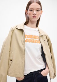 Beżowa, lekka kurtka z zamkiem z przodu, dużym kołnierzem i boczną kieszenią, noszona na białym T-shircie z pomarańczowym retro napisem. Ciemnogranatowe dżinsy.