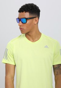 Tricou sportiv galben neon cu mâneci scurte, dungi gri contrastante pe umeri și un mic logo Adidas pe piept.