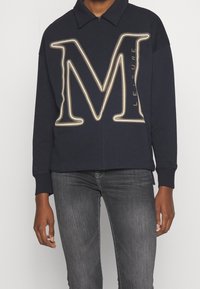 Felpa zip con cappuccio di colore blu navy, decorata con una grande lettera "M" a contorno dorato e la scritta "LEISURE". Presenta un colletto e polsini a costine.