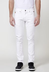 Jeans de algodón blancos con corte ajustado, diseño de cinco bolsillos, cierre con botón y un acabado texturizado sutil. Combinados con zapatillas negras.