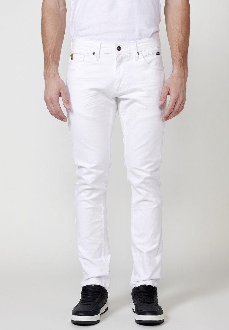 Jeans de algodón blancos con corte ajustado, diseño de cinco bolsillos, cierre con botón y un acabado texturizado sutil. Combinados con zapatillas negras.