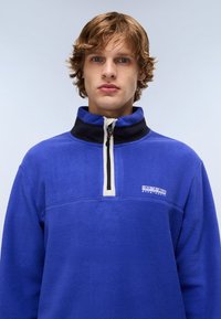 Blue fleece pullover with a half-zip collar, black trim, and cream accents. Features a front logo and textured fabric.