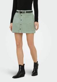 Femme portant une jupe boutonnée en velours côtelé vert clair avec poches avant, ceinture noire, haut à manches longues noir et bottines noires.