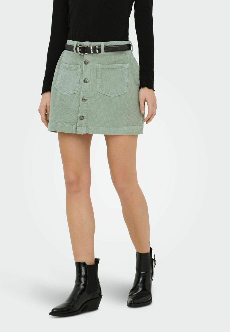 Femme portant une jupe boutonnée en velours côtelé vert clair avec poches avant, ceinture noire, haut à manches longues noir et bottines noires.