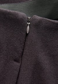 Ritssluiting detail op een donkergrijze stof, met een soepele textuur en een metalen ritsen trekker, met daarboven een groene geweven tape.