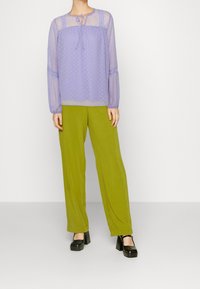 Blouse lilas transparente à pois avec un col à nouer, associée à un pantalon large vert et des talons compensés noirs.