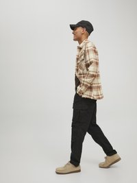 Braun-weiße karierte Flanellshirt, schwarze Cargohosen, beigefarbene Schnürschuhe und eine schwarze Mütze, getragen von einer Person im Profil.