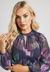 Genomskinlig blommönstrad blus i lila och gröna nyanser med nyckelhålsutskärning och volangärmar. Lätt, flödande tyg.