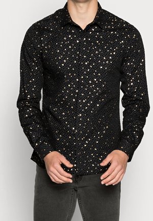 Homme portant une chemise noire à manches longues avec des pois dorés brillants et un pantalon gris foncé, les mains positionnées près de la taille.