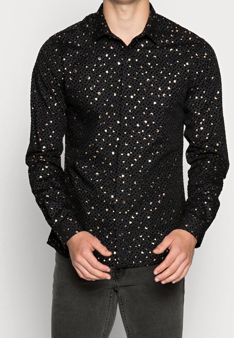 Homme portant une chemise noire à manches longues avec des pois dorés brillants et un pantalon gris foncé, les mains positionnées près de la taille.