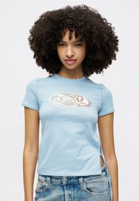 Camiseta azul clara de mangas curtas com um design ajustado, fendas laterais e um logótipo gráfico em castanho na parte da frente. Tecido suave e elástico.