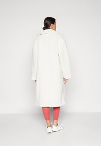 Cappotto bianco e morbido con un ampio colletto, spalle scese e design lunghezza ginocchio. L'indossatore ha indossato leggings rosa e sneakers bianche.