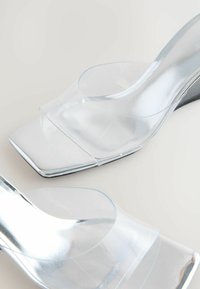Sandales à talons transparents en matériau clair avec un bout carré, un design ouvert et des accents métalliques sur le talon. Texture de surface lisse.