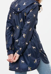 Marinblå vattentät regnkappa med huva, prydd med ett lekfullt mönster av hundar och människor som håller i paraplyer. Elastiska ärmslut och dragsko i midjan.