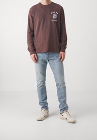 Camicia a maniche lunghe marrone con testo grafico bianco, abbinata a jeans azzurri chiari e sneakers crema con dettagli rossi.