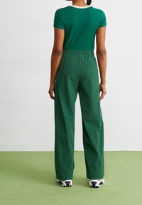 Persoon met gevlochten haar, met de rug naar de camera gekeerd, draagt een aangesloten groen T-shirt, losse groene broek met een elastische tailleband en witte sneakers met een zwarte zool.