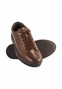 Brązowe skórzane sneakersy zapinane na sznurówki z teksturowaną gumową podeszwą, jeden but stoi pionowo, a drugi pokazuje wzór przyczepności podeszwy za nim.