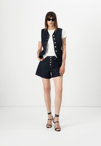Denimvest met zilveren knopen, gedragen over een wit T-shirt, gecombineerd met donkere denim shorts en zwarte hakken met straps. Fitted ontwerp, casual stijl.