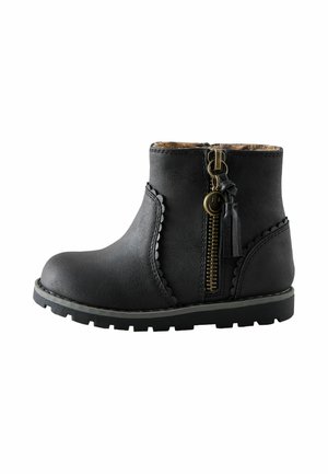 Sort ankelstøvle med bølget kant, sides lynlås med smiley charm og tassel, og en robust sål på en hvid baggrund.