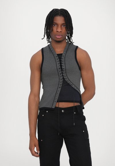 Gilet gris ajusté avec un devant à lacets, un col rond et des accents noirs. Porté sur un débardeur bleu marine, associé à un pantalon cargo noir.