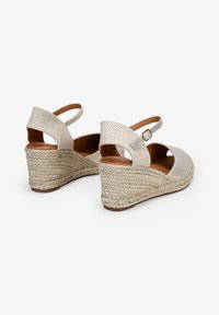 Sandalias de cuña con suela de yute trenzada, parte superior de textil metálico beige, talón abierto y correa al tobillo con una hebilla decorativa.