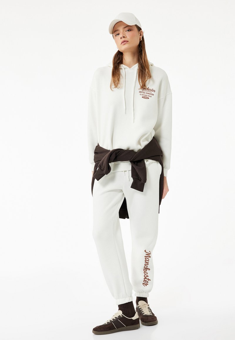 Sudadera blanca con texto marrón "Manchester", combinada con pantalones de chándal blancos a juego. El modelo lleva un suéter marrón atado a la cintura y una gorra blanca.