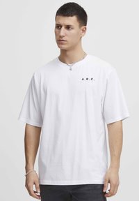 Hvid bomulds T-shirt med korte ærmer og en afslappet pasform. Har et sort "A.R.C." print på venstre bryst. Glat tekstur.