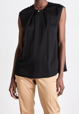 Personne portant un chemisier noir sans manches avec un détail pli boutonné, associé à un pantalon beige sur mesure, sur un fond uni.