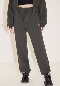 Dunkelgrüne, gerippte Jogginghosen mit elastischem Bund und Kordelzug; verfügen über seitliche Schlitze und ein weißes Logo auf der linken Hüfte; kombiniert mit schwarzen Sneakers.
