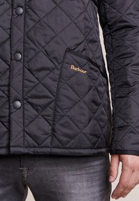 Casaco acolchoado preto com padrão de losangos, botões de pressão, bolso lateral e logótipo "Barbour" bordado em dourado. Tecido macio e texturizado.