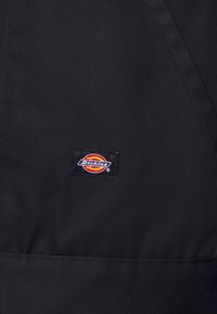 Svart tyg med en slät textur som har ett Dickies-logotypmärke i röd, gul och blå i mitten. Synliga sömmar.