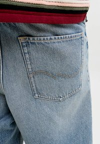 Tasca posteriore di jeans in denim azzurro chiaro con cuciture curve, indossati con un maglione a righe infilato in vita.