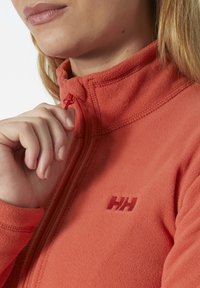 Veste en polaire orange avec col haut, dotée d'une fermeture éclair et d'un logo ton sur ton sur la poitrine. Texture lisse et coupe ajustée.